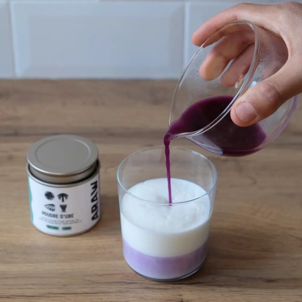 Une main verse du lait d’ube violet dans un verre de lait, à côté d’une boîte de poudre Araw
