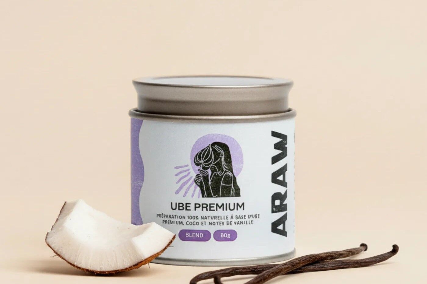 Une boîte de poudre d’ube Araw La Blend posée entre un morceau de noix de coco et des gousses de vanille, sur fond beige