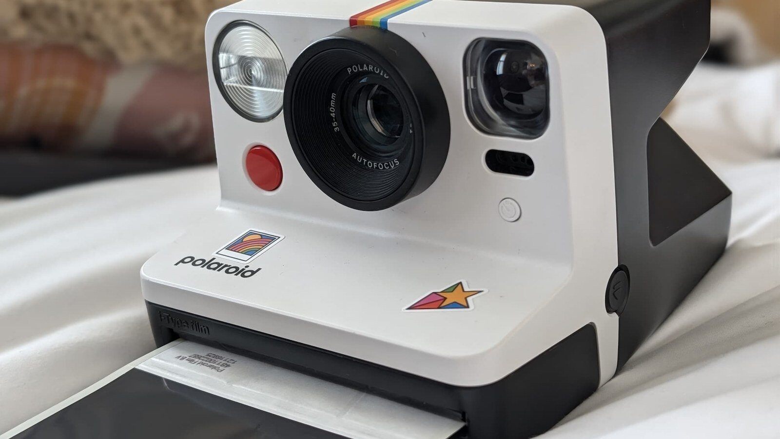 Appareil photo instantané Polaroid Now Génération 2 blanc posé sur un lit, vu de trois quarts, une photo sortant de la fente