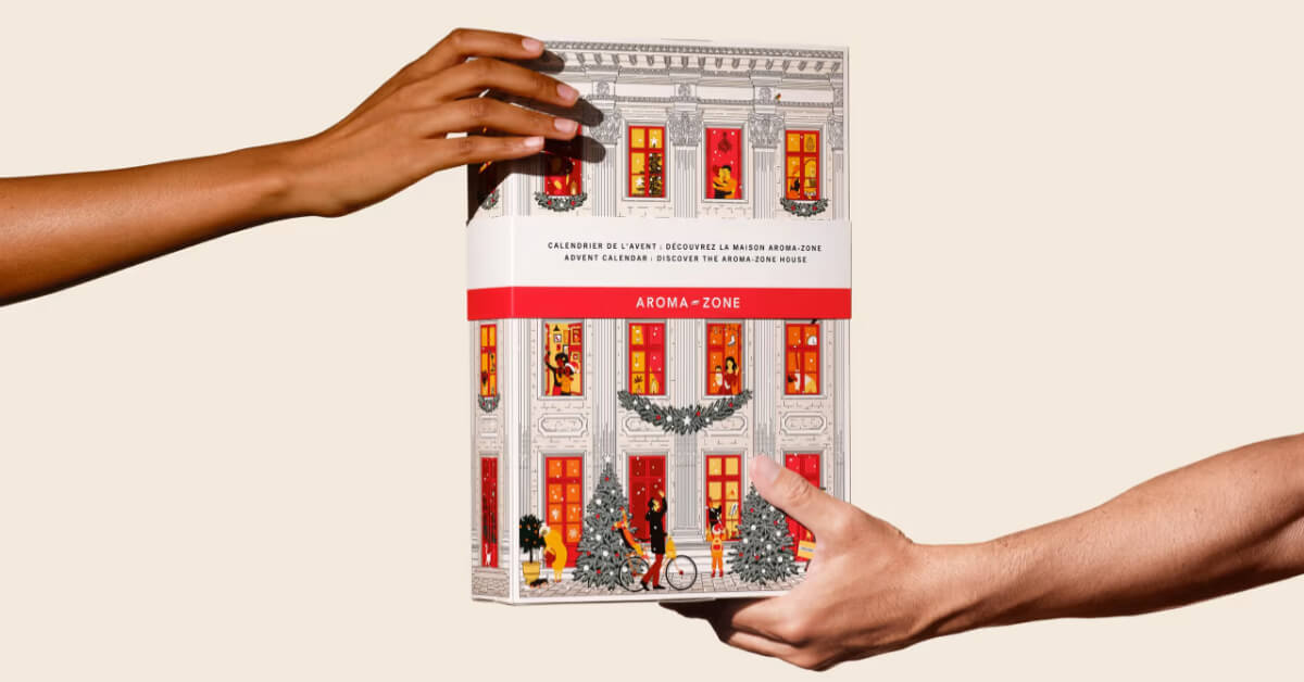 Le calendrier de l’Avent Aroma-Zone 2026 tenu par deux mains : une façade d’immeuble blanche aux fenêtres rouges illuminées, avec un bandeau « Découvrez la maison Aroma-Zone »