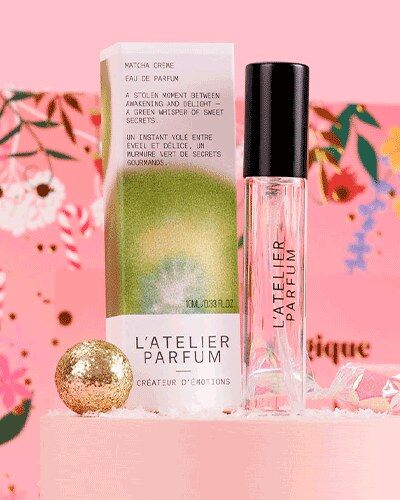 Une eau de parfum L’Atelier Parfum et son étui vert posés devant le décor fleuri de la papillote Glowria