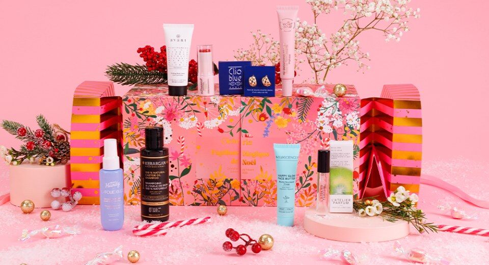 Le calendrier de l’Avent Glowria 2026 en forme de papillote géante rose et dorée, entouré de produits de beauté dont un shampooing Kerargan, une brume Merci Handy et une boîte Clio Blue
