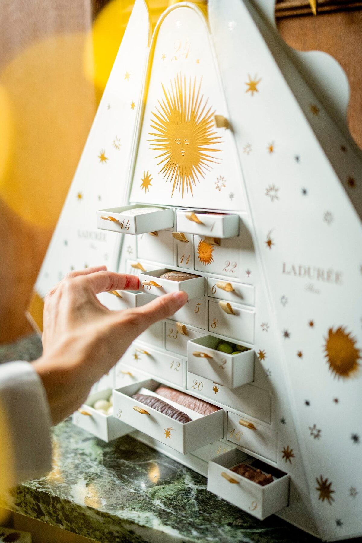 Une main ouvre l’un des tiroirs du calendrier de l’Avent Ladurée 2026, décoré d’un soleil doré