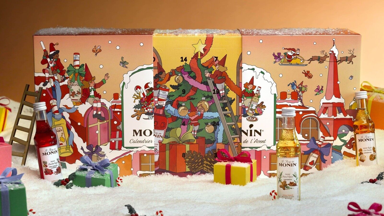 Calendrier de l’Avent Monin illustré de lutins et d’un sapin, posé dans la neige entre des cadeaux et des mini-bouteilles de sirop