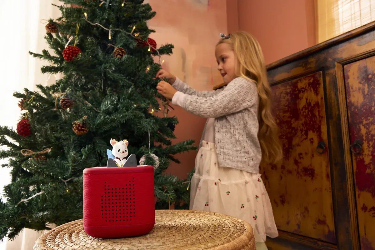 Une petite fille décore un sapin de Noël, à côté d’une Toniebox rouge surmontée d’une figurine