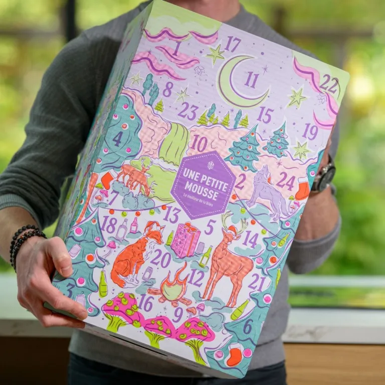 Un homme tient le calendrier de l’Avent bières Une Petite Mousse 2026, un grand carton illustré d’une forêt enchantée aux tons pastel avec des cases numérotées