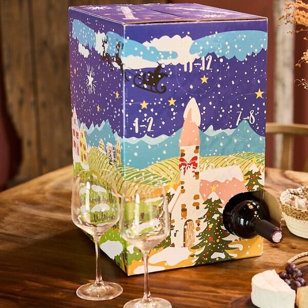 Le calendrier de l’Avent du vin de La Boîte du Fromager, un grand carton illustré d’un village enneigé, posé sur une table avec deux verres et une bouteille