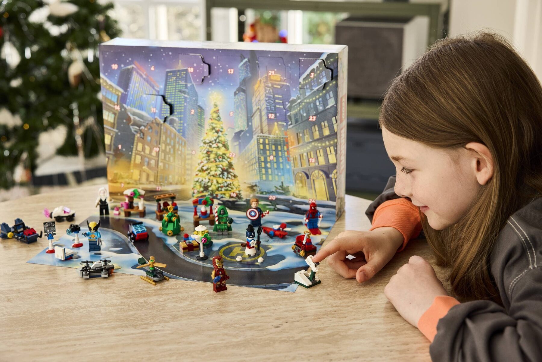 Une enfant joue avec les minifigurines et petits véhicules du calendrier de l’Avent LEGO Marvel 2026, devant la boîte illustrée d’une ville et d’un sapin de Noël