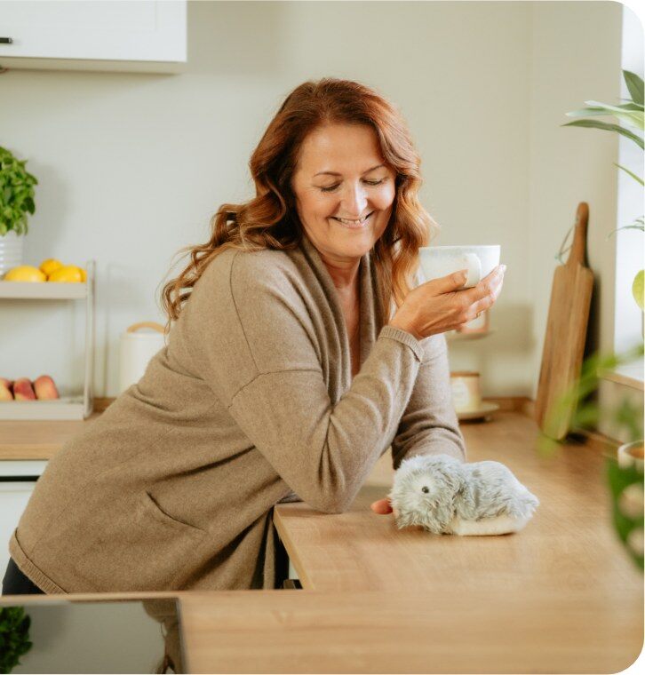 Une femme sourit en tenant une tasse, un Moflin argent posé sur le plan de travail de la cuisine