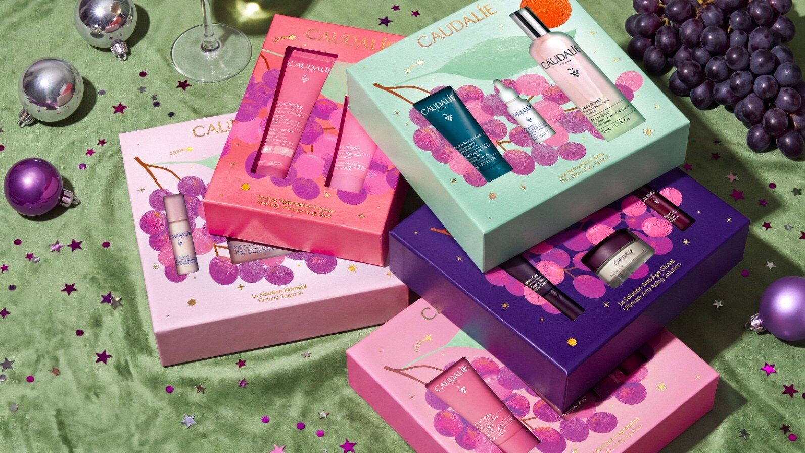 Coffrets de Noël Caudalie 2026 roses, violets et vert d’eau illustrés de grappes de raisin, posés sur un velours vert avec boules de Noël