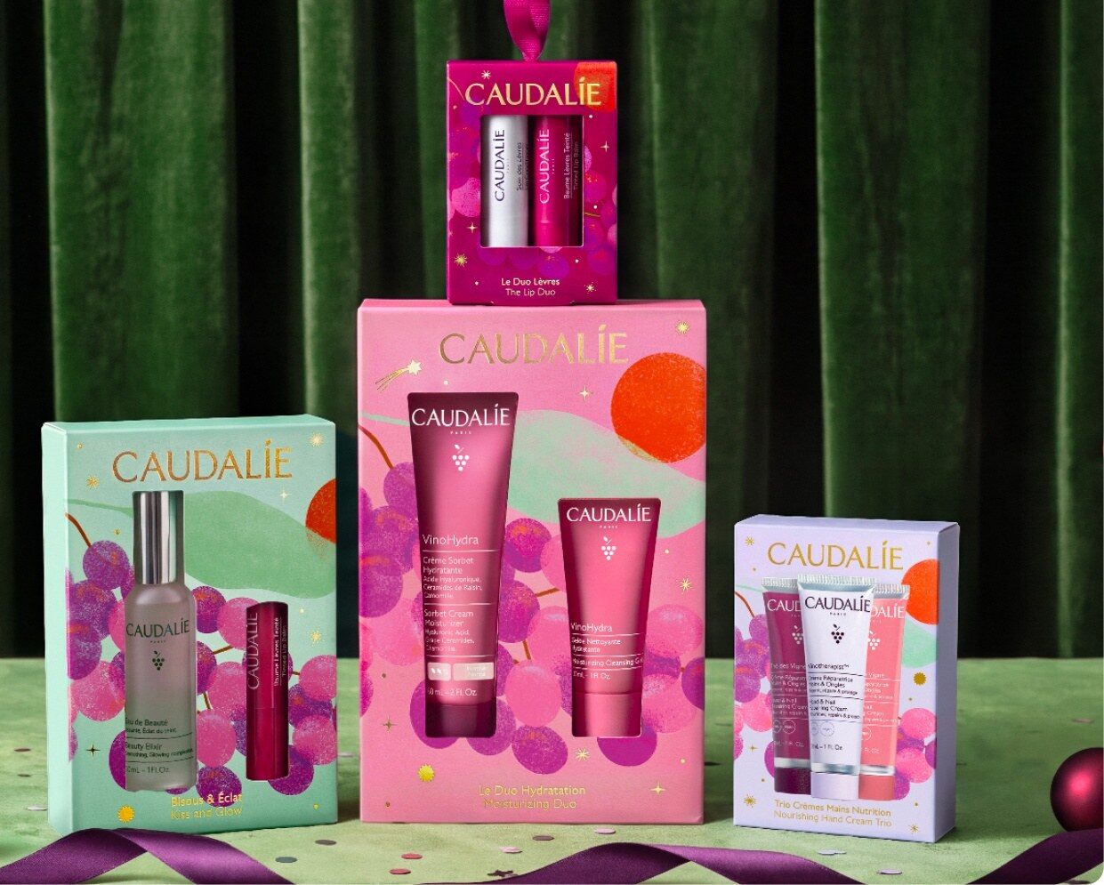 Coffrets de Noël Caudalie 2026 VinoHydra, Eau de Beauté et duo lèvres posés devant un rideau de velours vert