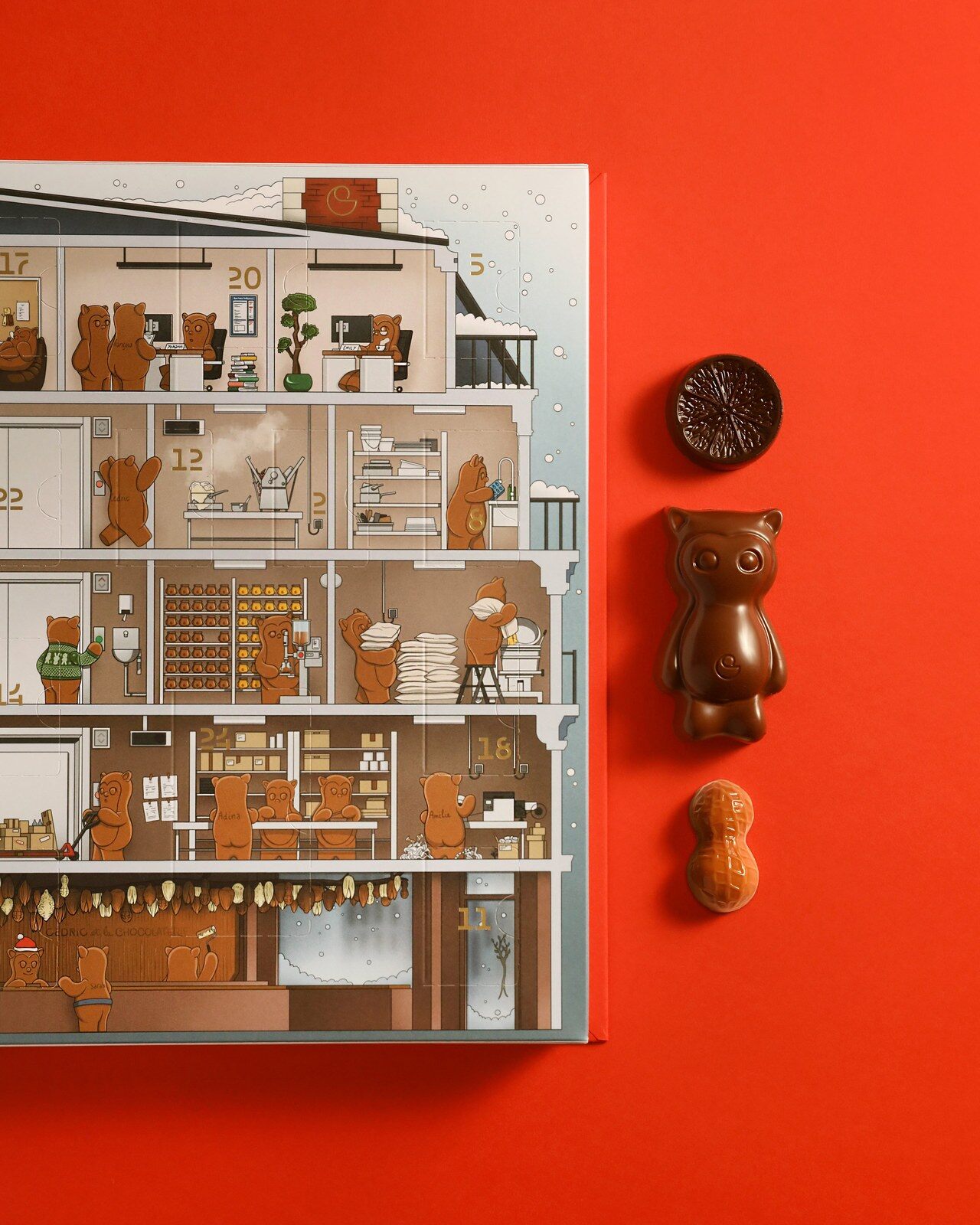 Le calendrier de l’Avent Tome 2 de Cédric et la Chocolaterie ouvert sur fond rouge, à côté d’un chocolat en forme d’agrume, d’un ourson en chocolat et d’une cacahuète en chocolat