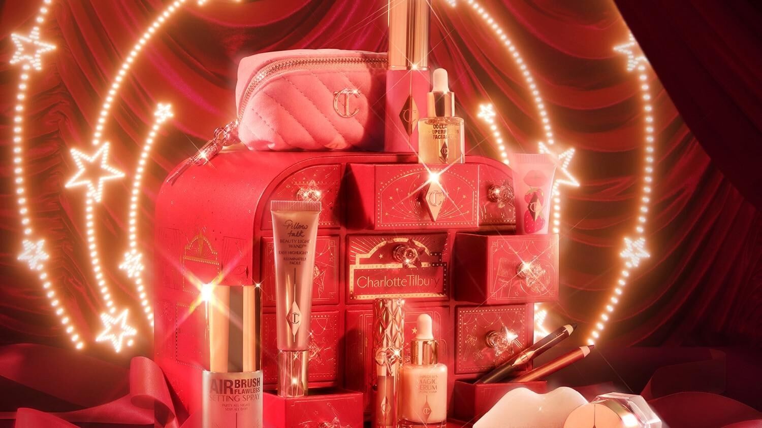 Le calendrier de l’Avent Charlotte Tilbury 2026 rouge et or, ses produits posés autour, devant un rideau rouge et une arche d’étoiles lumineuses