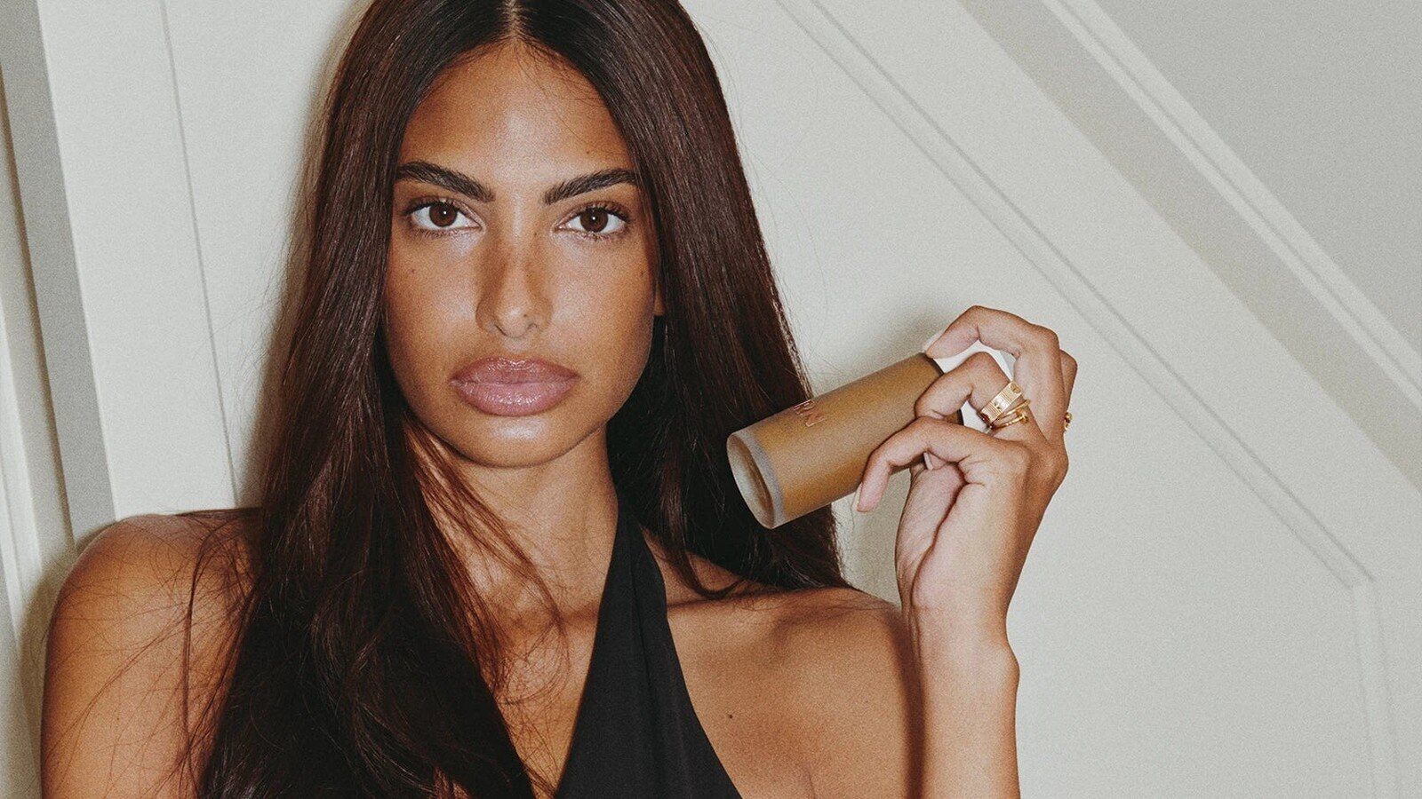 Jeune femme brune tenant le flacon du Milk Teinté Charly Salvator x FIRN devant un mur clair