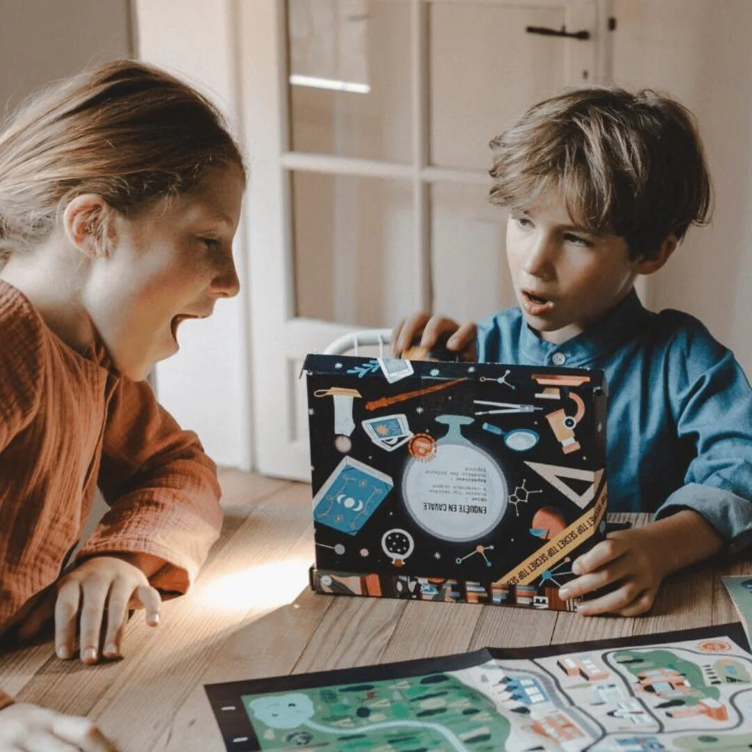 Deux enfants ouvrent avec enthousiasme une box En Cavale illustrée d’objets scientifiques, posée sur une table à côté d’une carte