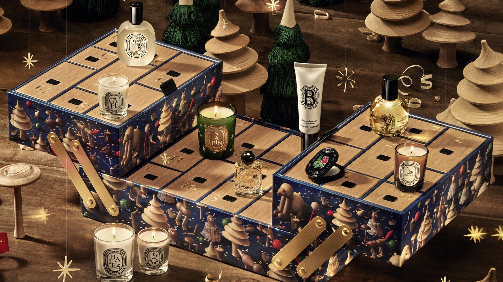 Le calendrier de l’Avent Diptyque 2026 ouvert, bougies et flacons posés sur ses tiroirs, au milieu de sapins en bois tourné