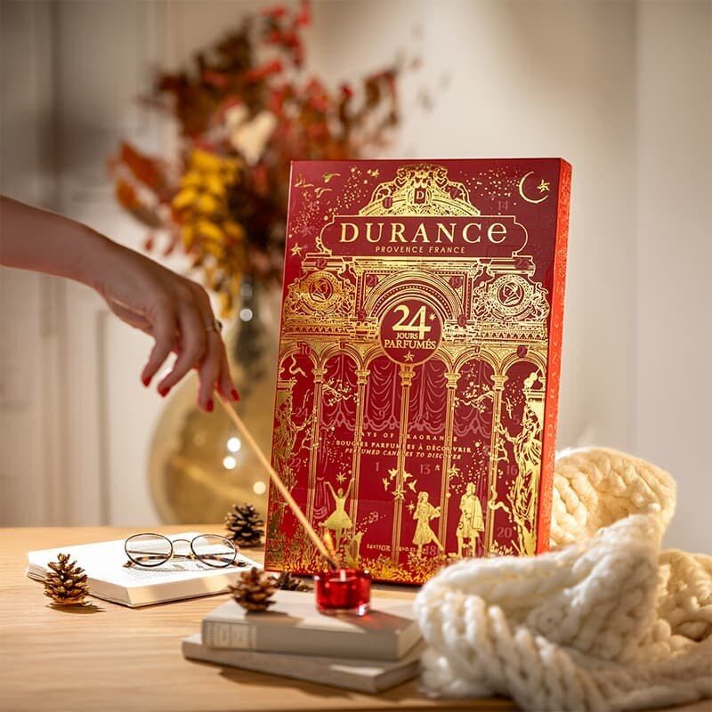 Une main allume une bougie devant le calendrier de l’Avent Durance 2026 posé sur une table, près d’un plaid en grosse maille et de pommes de pin