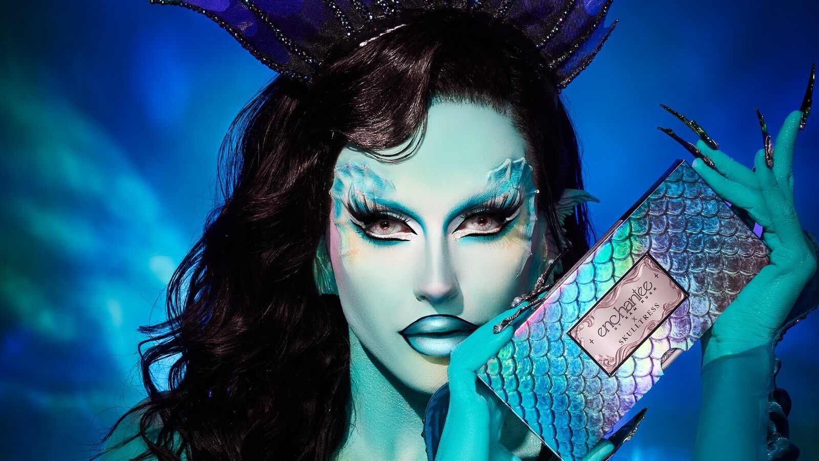 Une artiste maquillée en sirène bleu-vert, couronne d’écailles sur la tête, tient la palette irisée Dark Siren contre son visage