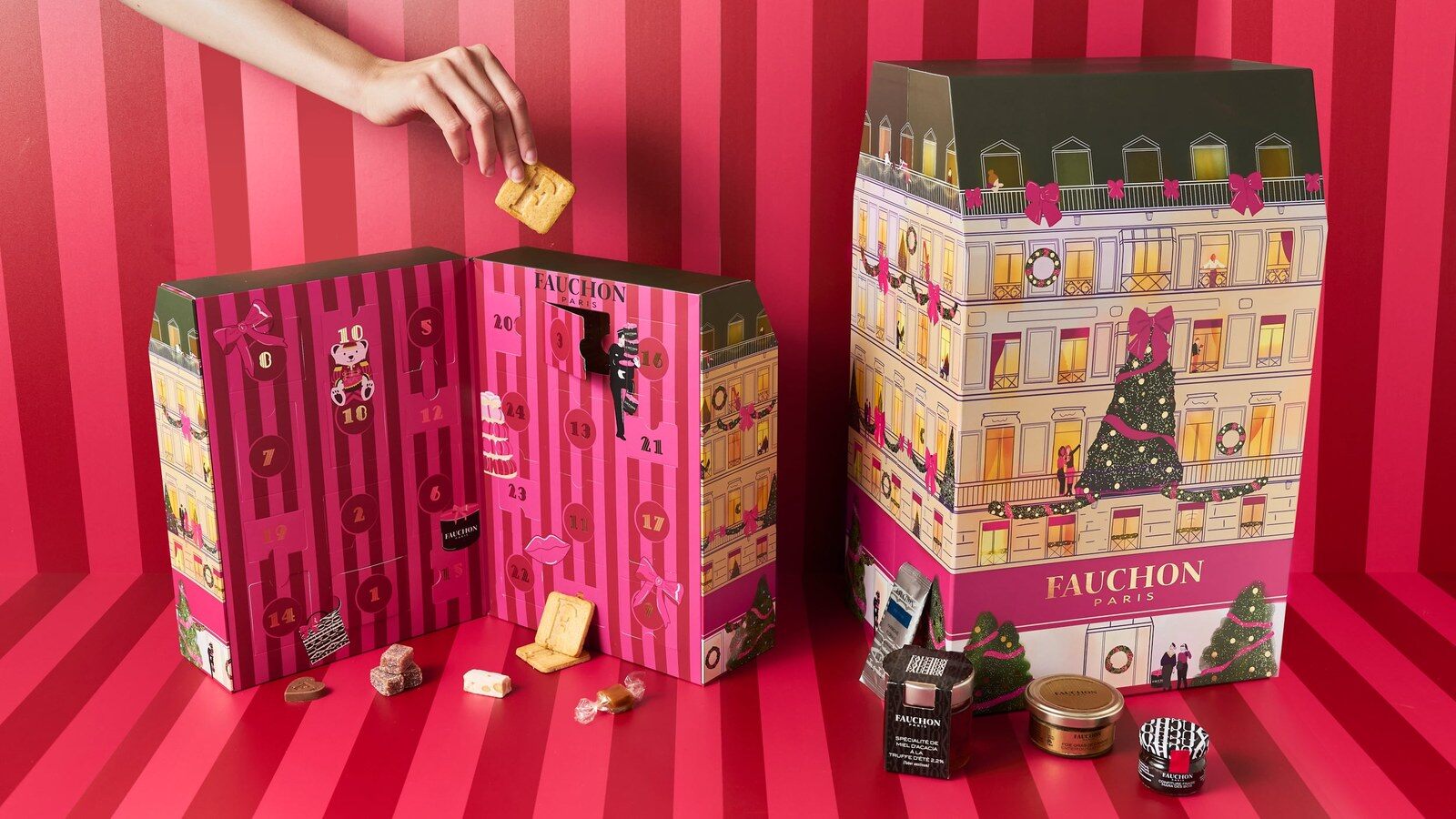 Une main pioche un biscuit dans le calendrier de l’Avent confiserie Fauchon ouvert, à côté du calendrier Luxe en forme d’immeuble parisien