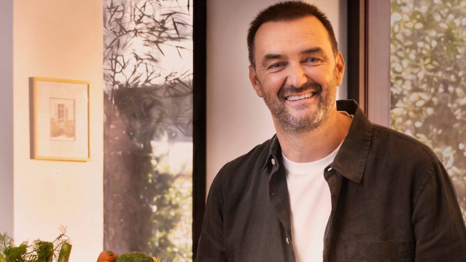 Cyril Lignac souriant dans une cuisine lumineuse, avec des légumes frais au premier plan