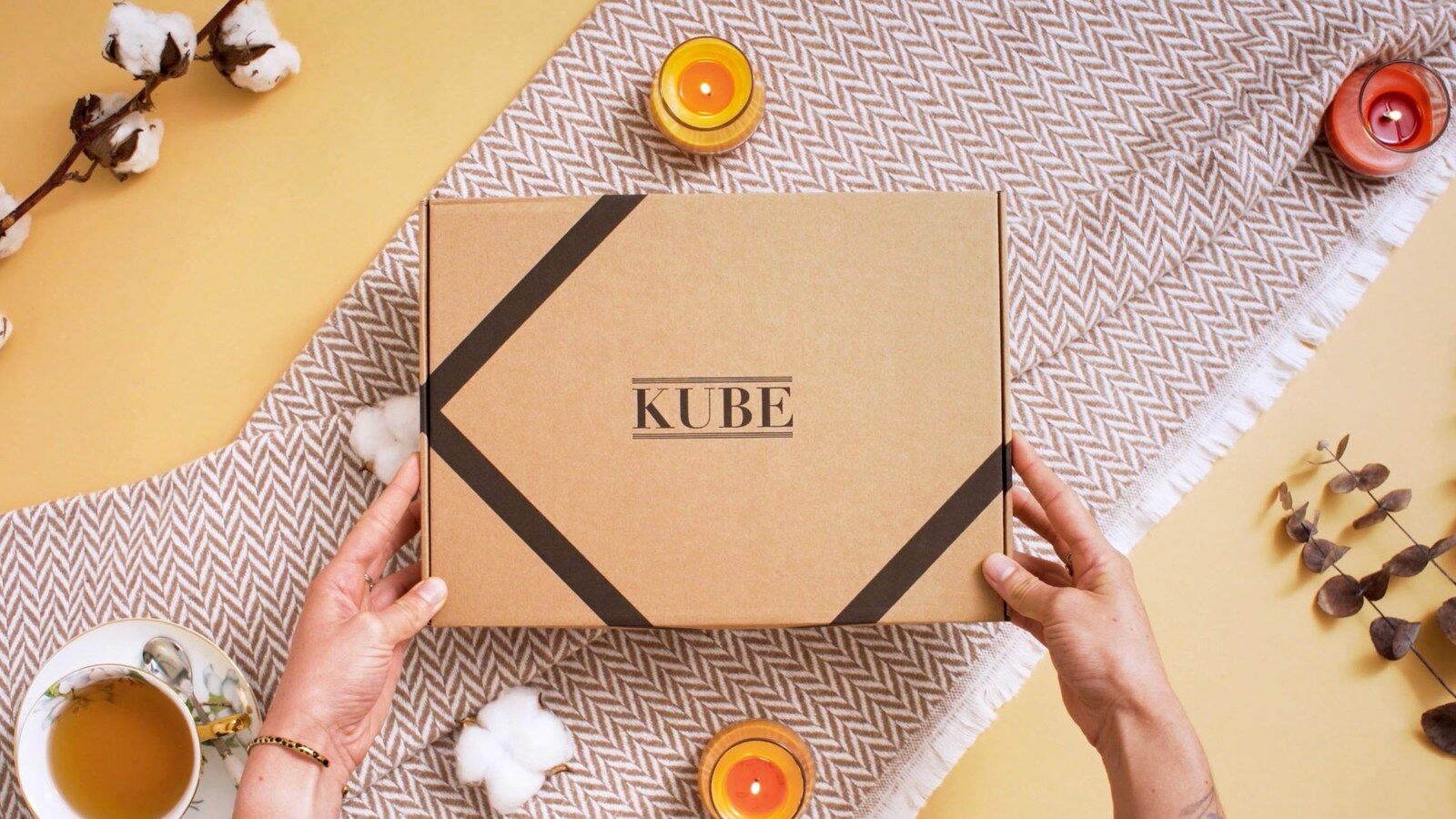 Mains tenant une box livre Kube fermée sur un plaid, à côté d’une tasse de thé, de bougies et d’eucalyptus