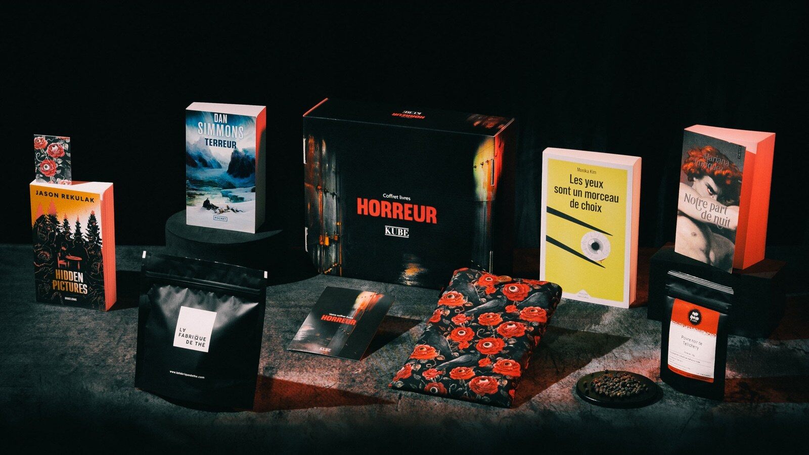 Le Coffret livres Horreur de Kube entouré de ses quatre romans, d’un étui à livre fleuri et d’un sachet de thé, dans une ambiance sombre