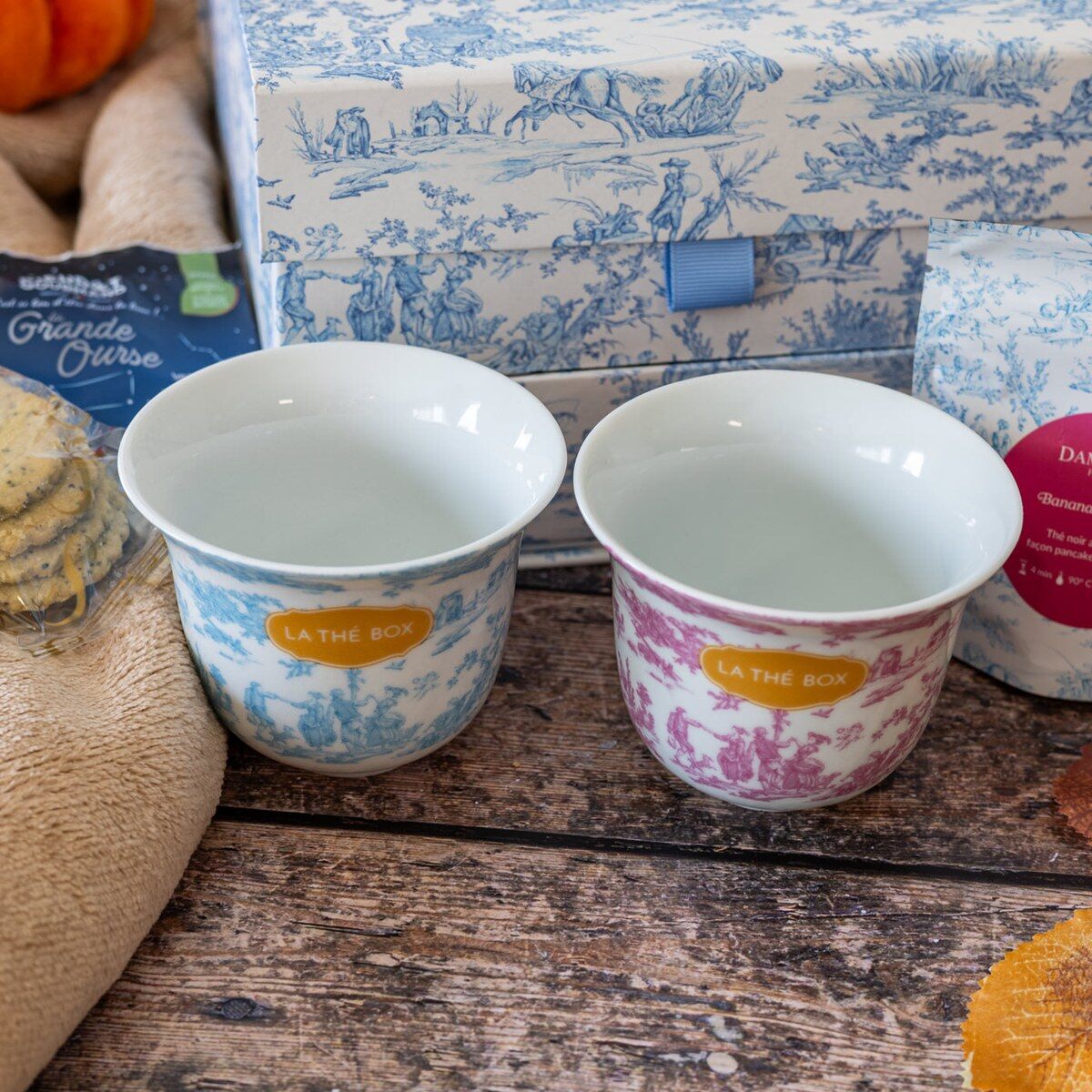 Deux tasses en toile de Jouy, une bleue et une rose, posées devant le coffret Maison de Famille