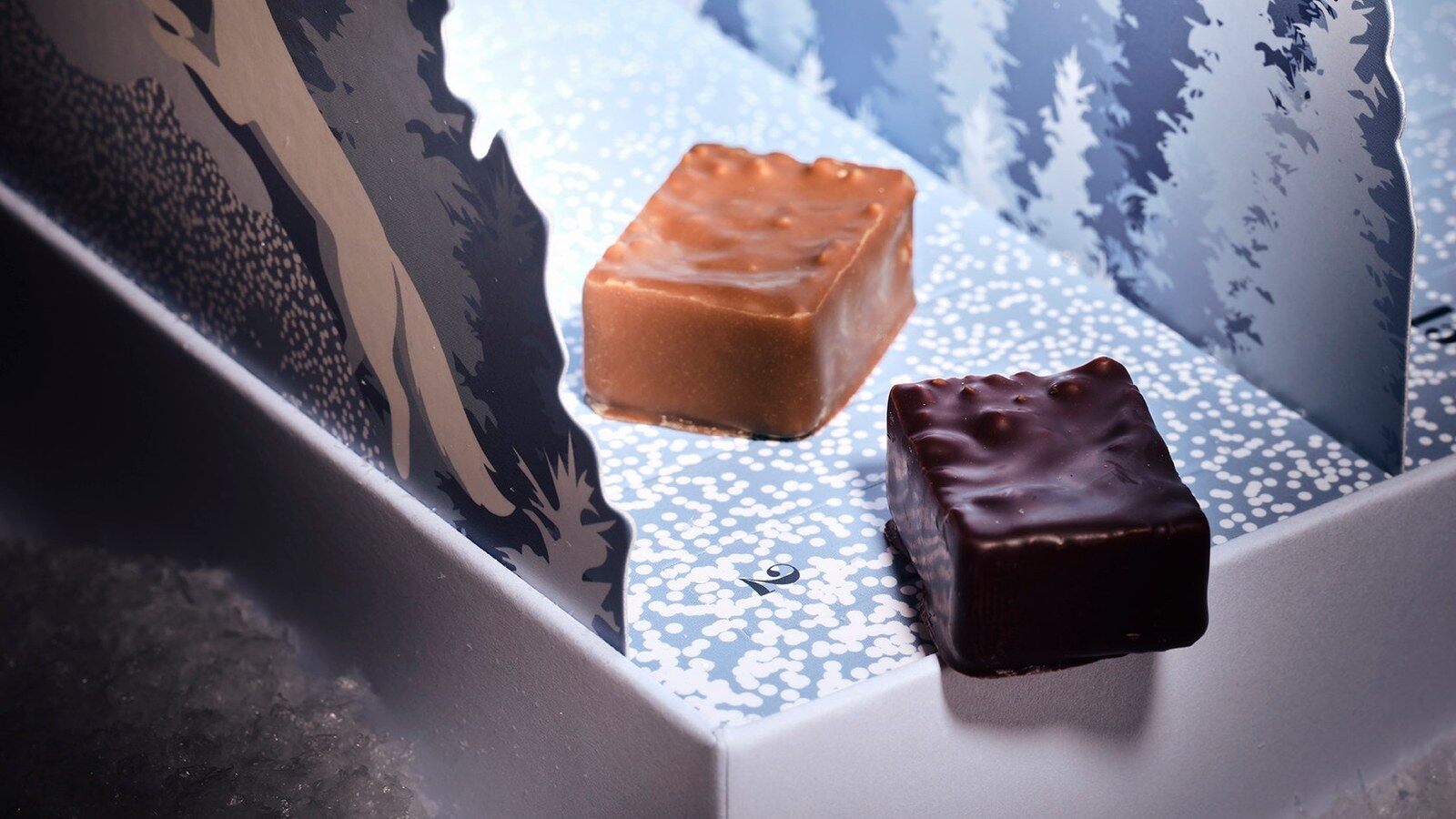 Deux bonbons de chocolat Lenôtre, au lait et noir, posés dans le calendrier de l’Avent ouvert au décor de forêt enneigée