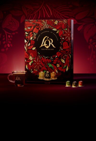 Le calendrier de l’Avent L’Or Espresso 2026 posé à côté d’une tasse de café et de capsules, sur un décor rouge
