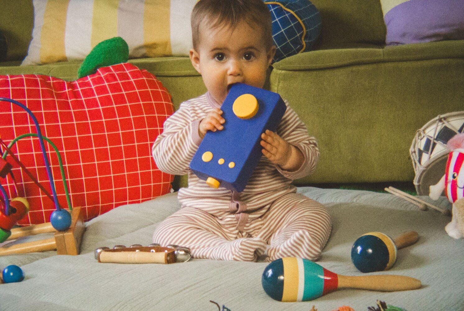 Bébé assis sur un tapis de salon, Ma 1ère Lunii bleue à la bouche, entouré de jouets en bois