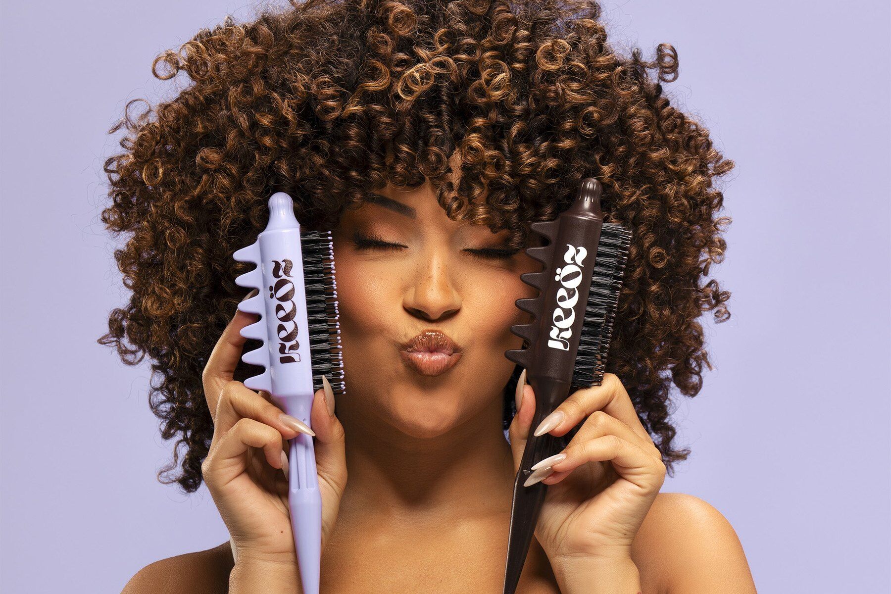 Femme aux cheveux bouclés tenant deux brosses Finger Coils Maison Keeöz, lilas et marron, sur fond lilas