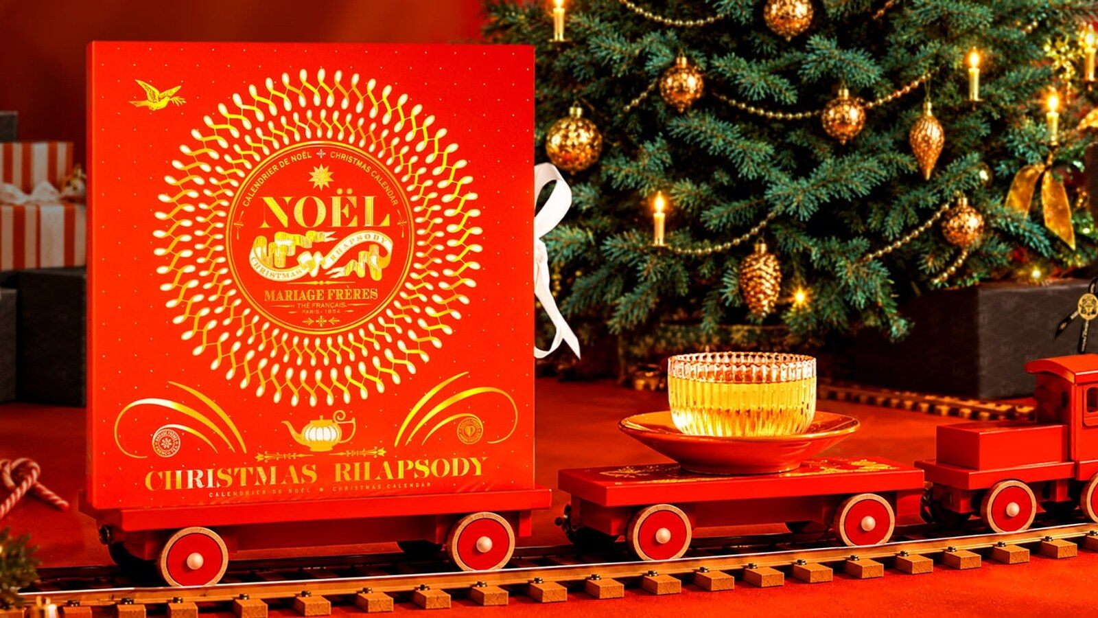 Le calendrier de l’Avent Mariage Frères Christmas Rhapsody 2026, écrin orange et or, posé sur un petit train de Noël rouge à côté d’une tasse de thé, devant un sapin illuminé
