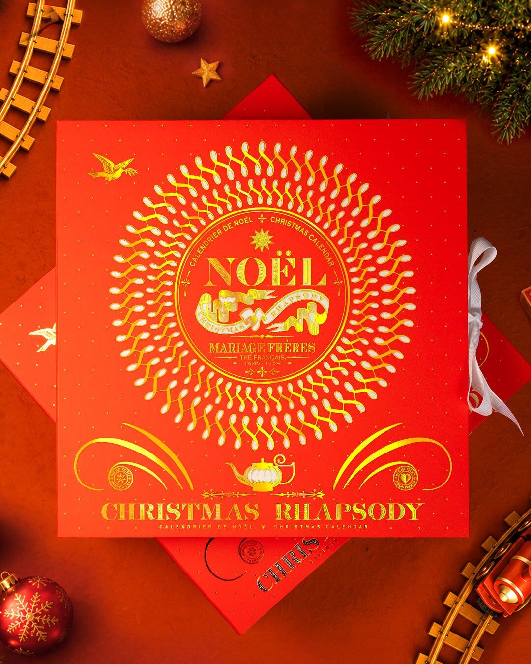 Le calendrier Christmas Rhapsody de Mariage Frères posé sur un petit train de Noël rouge devant un sapin illuminé