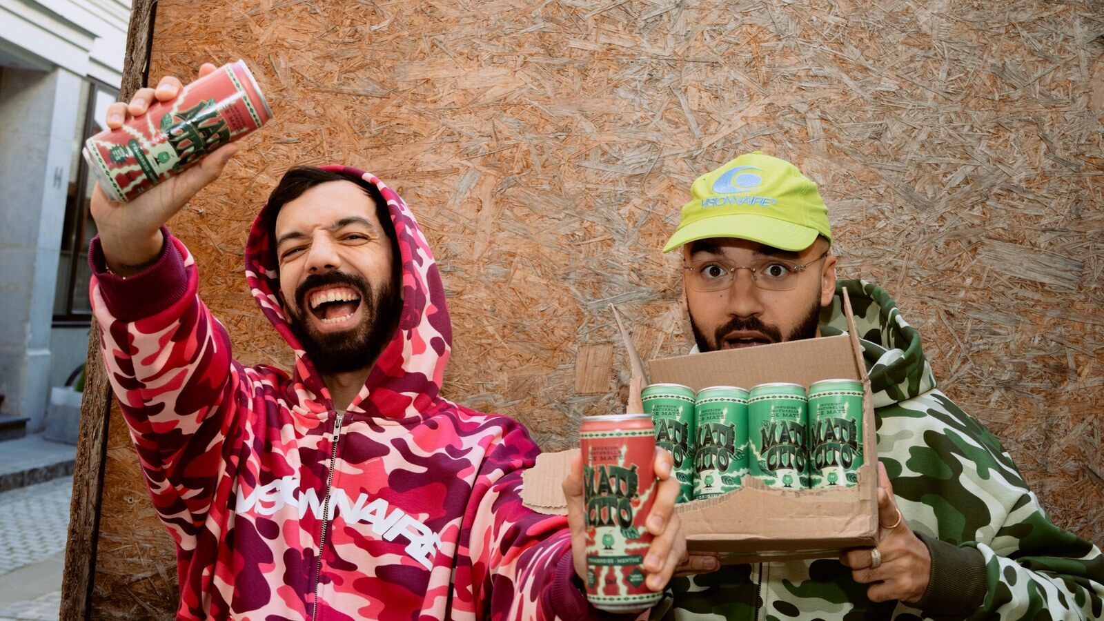 Bigflo et Oli brandissent des canettes Matécito et un carton de canettes vertes devant une palissade en bois