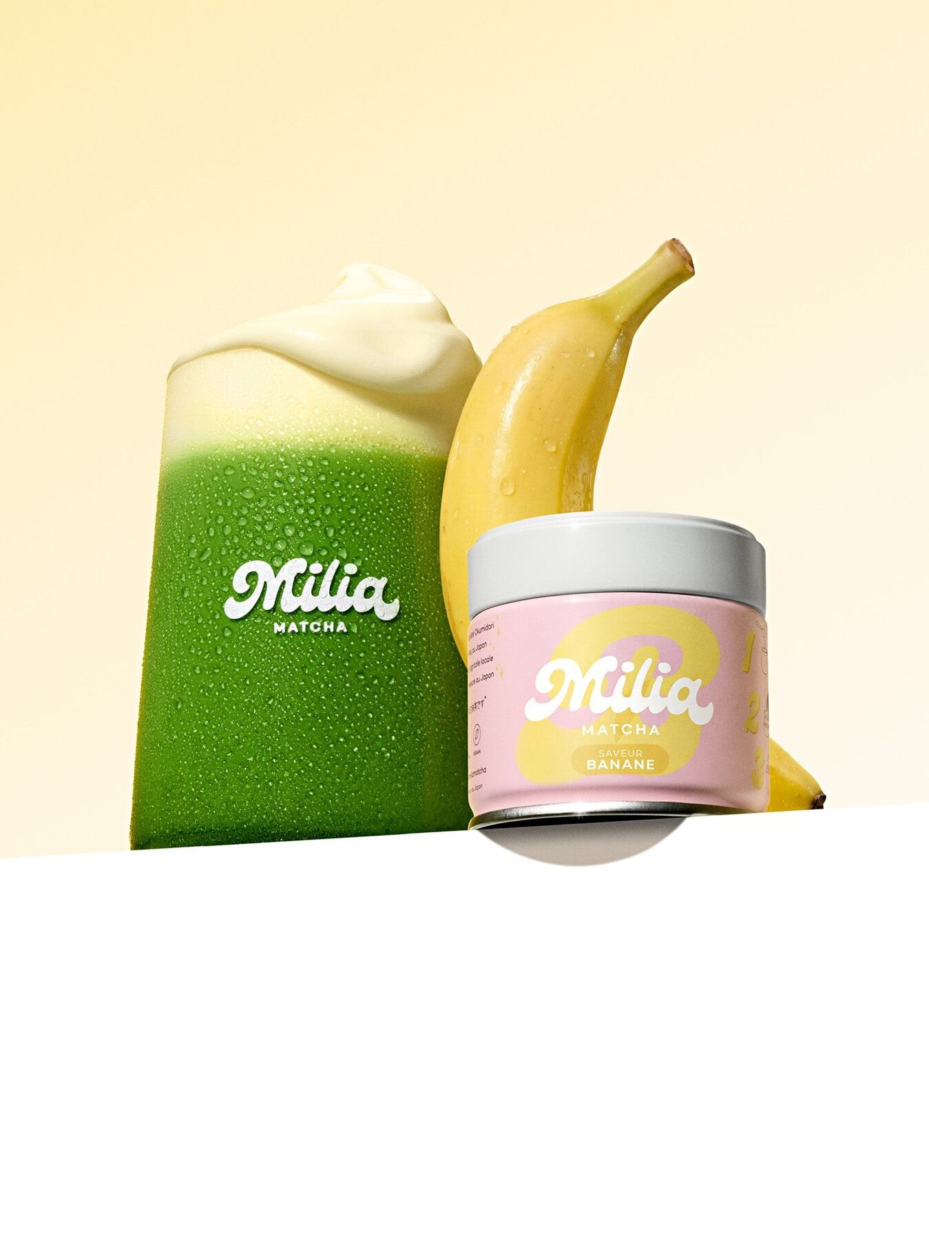 Verre de matcha latte vert surmonté de mousse, à côté d’une canette Milia Matcha Banane et d’une banane