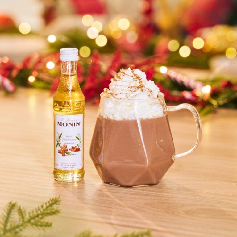 Chocolat chaud au sirop de pain d’épices Monin surmonté de crème, à côté d’une mini-bouteille