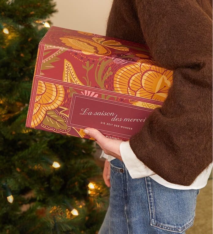 Une femme porte la Double Box My Little Box « La Saison des merveilles » sous le bras, devant un sapin de Noël