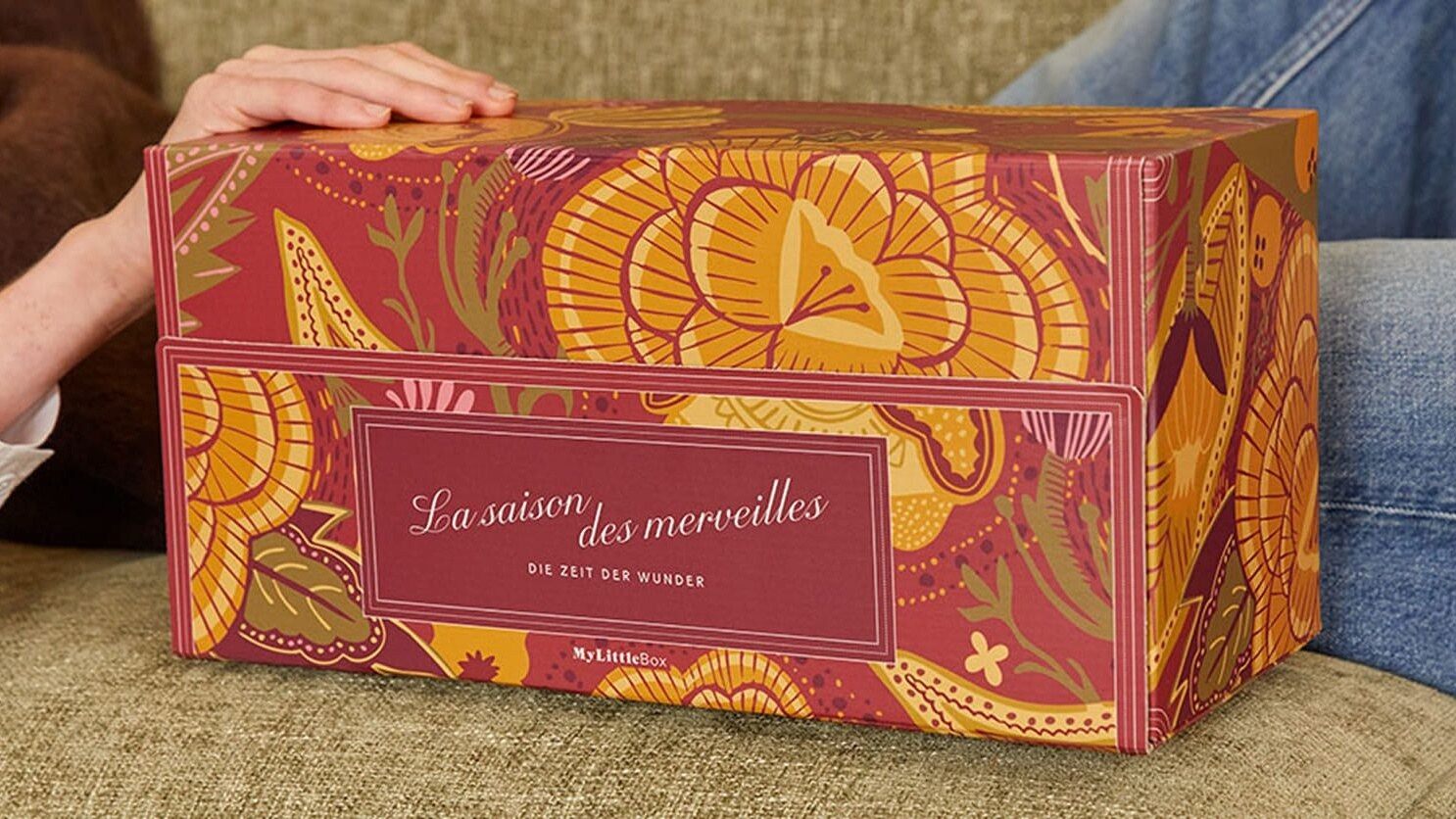 La Double Box de Noël My Little Box « La Saison des merveilles » posée sur un canapé, une main sur le couvercle