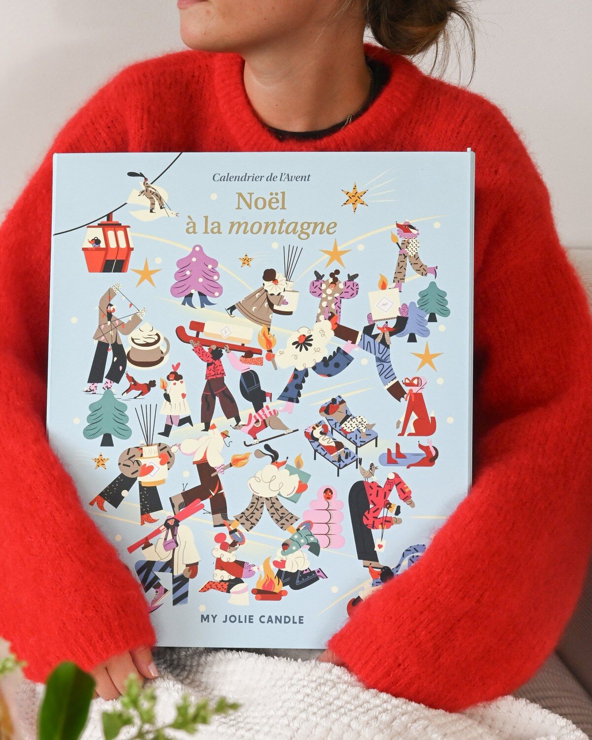 Une femme en pull rouge tient le calendrier de l’Avent My Jolie Candle 2026 Noël à la montagne