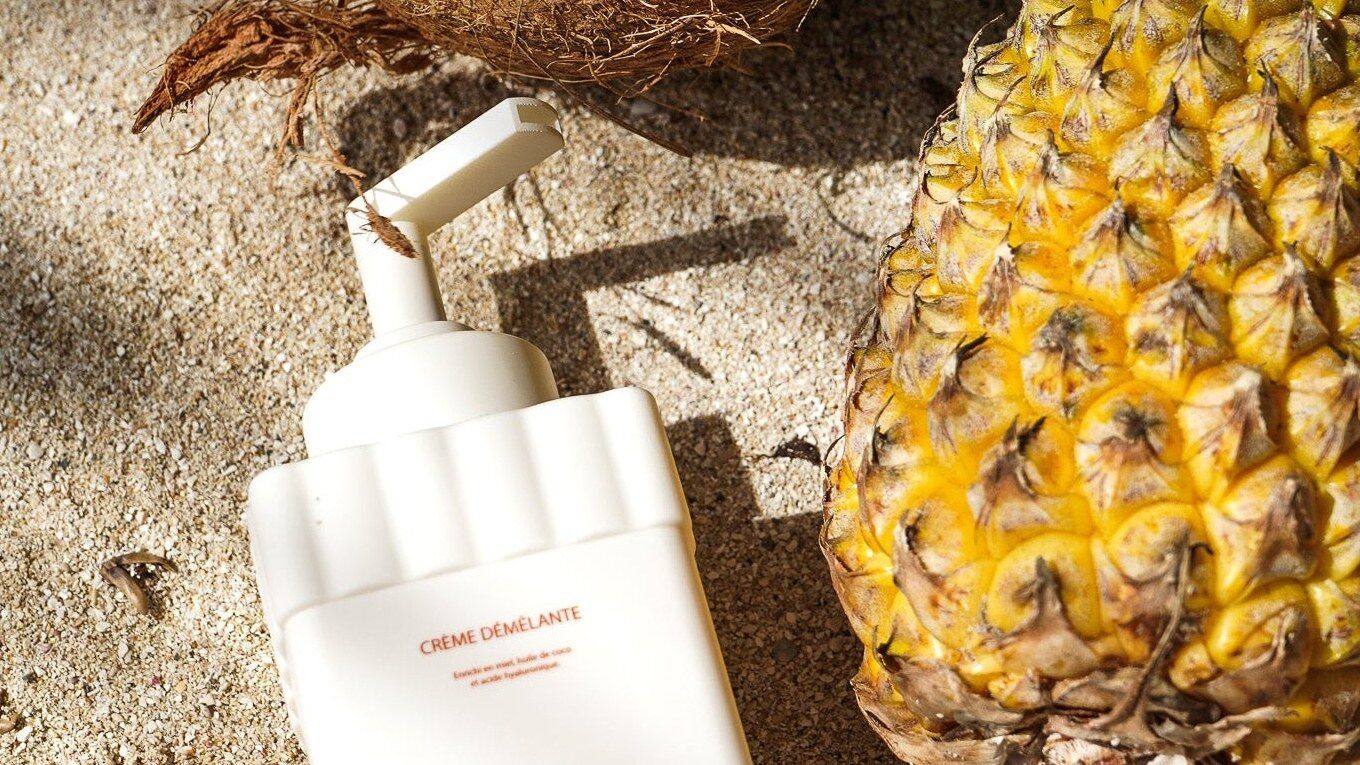 Le flacon blanc de crème démêlante Nurly posé sur le sable à côté d’un ananas