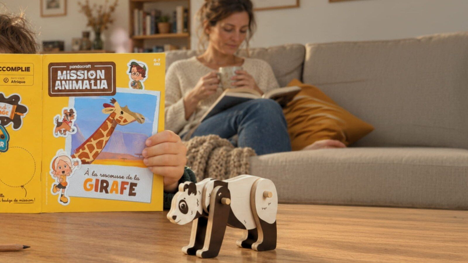 Un enfant lit un carnet Mission Animalia sur la table du salon, devant une figurine panda, sa mère sur le canapé