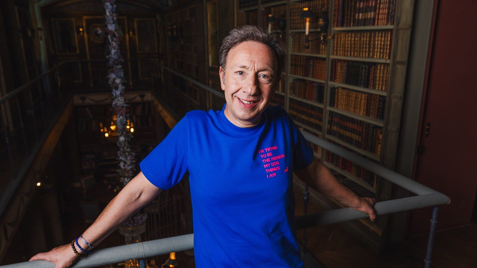 Stéphane Bern en t-shirt bleu, accoudé à la rambarde d’une bibliothèque de château