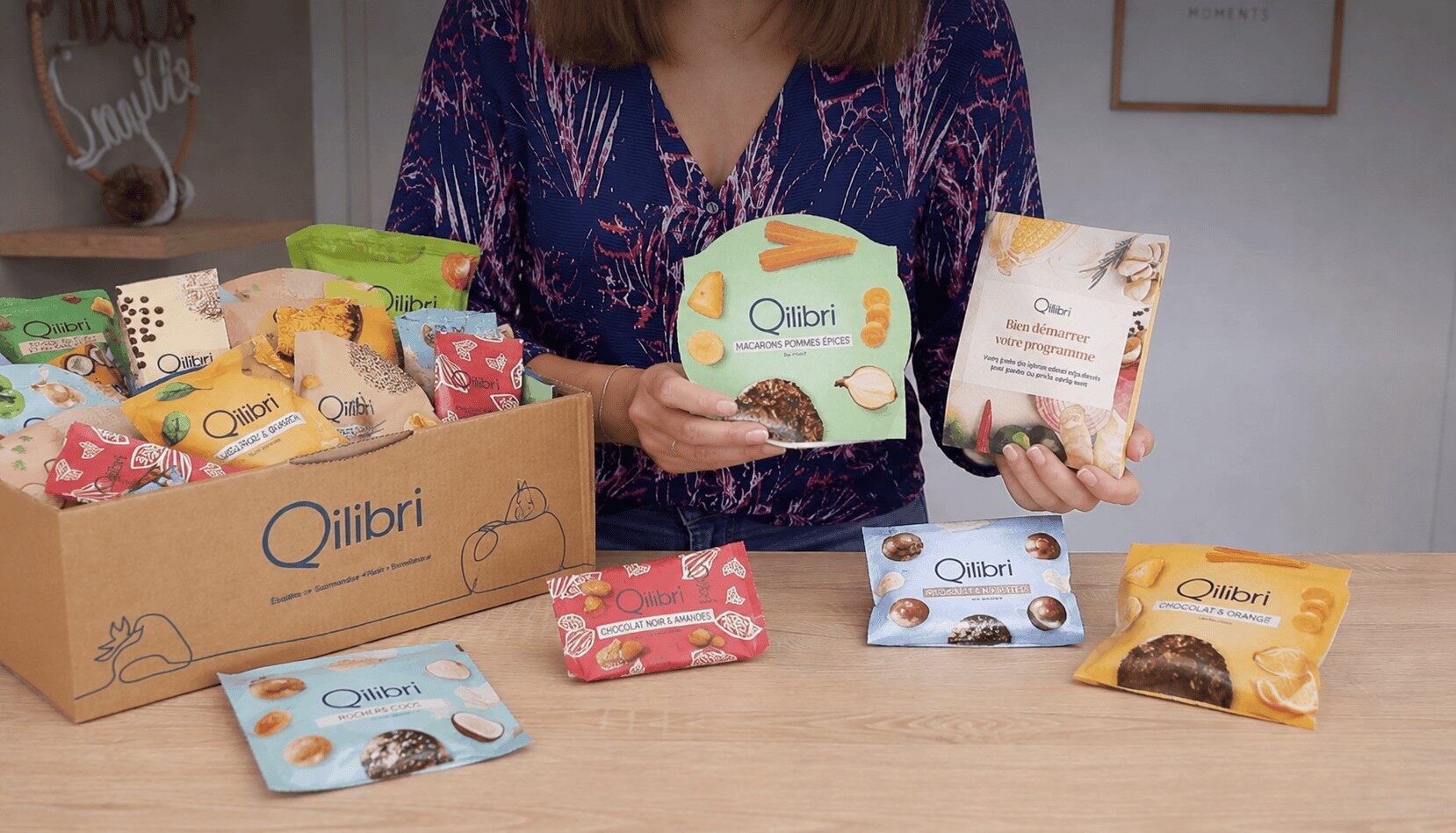 Une femme présente des sachets de collations Qilibri devant un colis de la marque ouvert sur une table