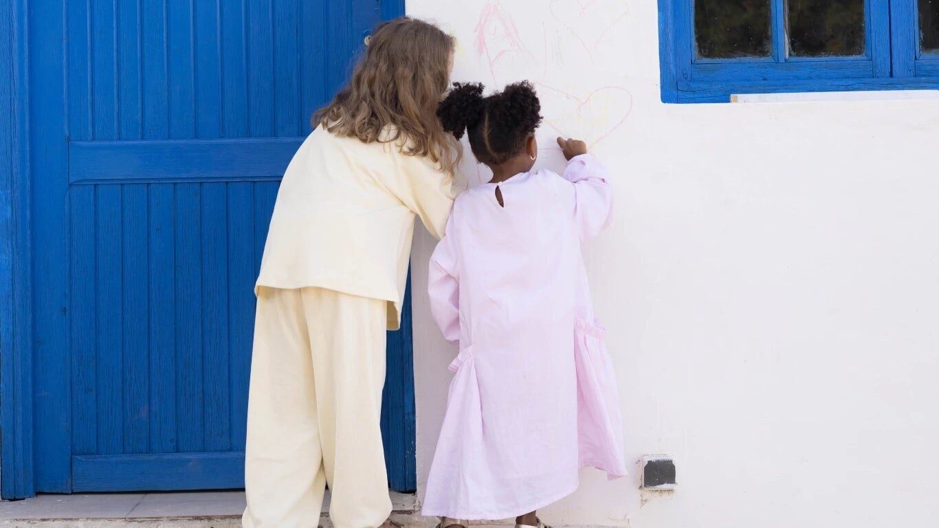 Deux enfants de dos dessinent sur un mur blanc près d’une porte bleue, en Robe Rosée et Ensemble Aube RAÏSHA