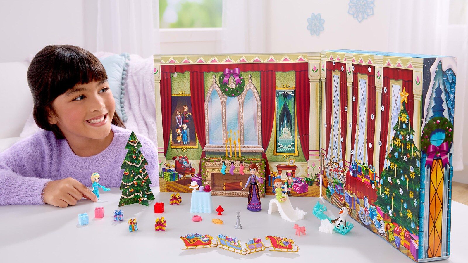 Une enfant souriante joue avec les poupées Anna et Elsa enfants devant le calendrier de l’Avent La Reine des Neiges 2026 déplié en salon de château