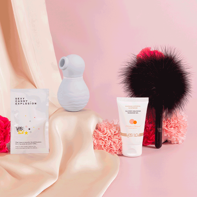 Flat-lay officiel de la Box Coquine : le stimulateur clitoridien, le plumeau noir, le sachet Sexy Candy Explosion et le tube de gel de massage YesForLov, disposés sur un drapé beige et rose.