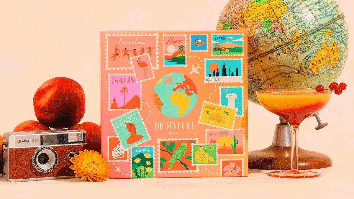 Visuel officiel de la Biotyfull Box d’octobre 2026 « Voyage autour du Monde » : la box orange ornée de vignettes de destinations, entourée d’un globe terrestre, d’un appareil photo et de fruits. Le visuel illustre le thème et la box, pas les produits qui la composent.