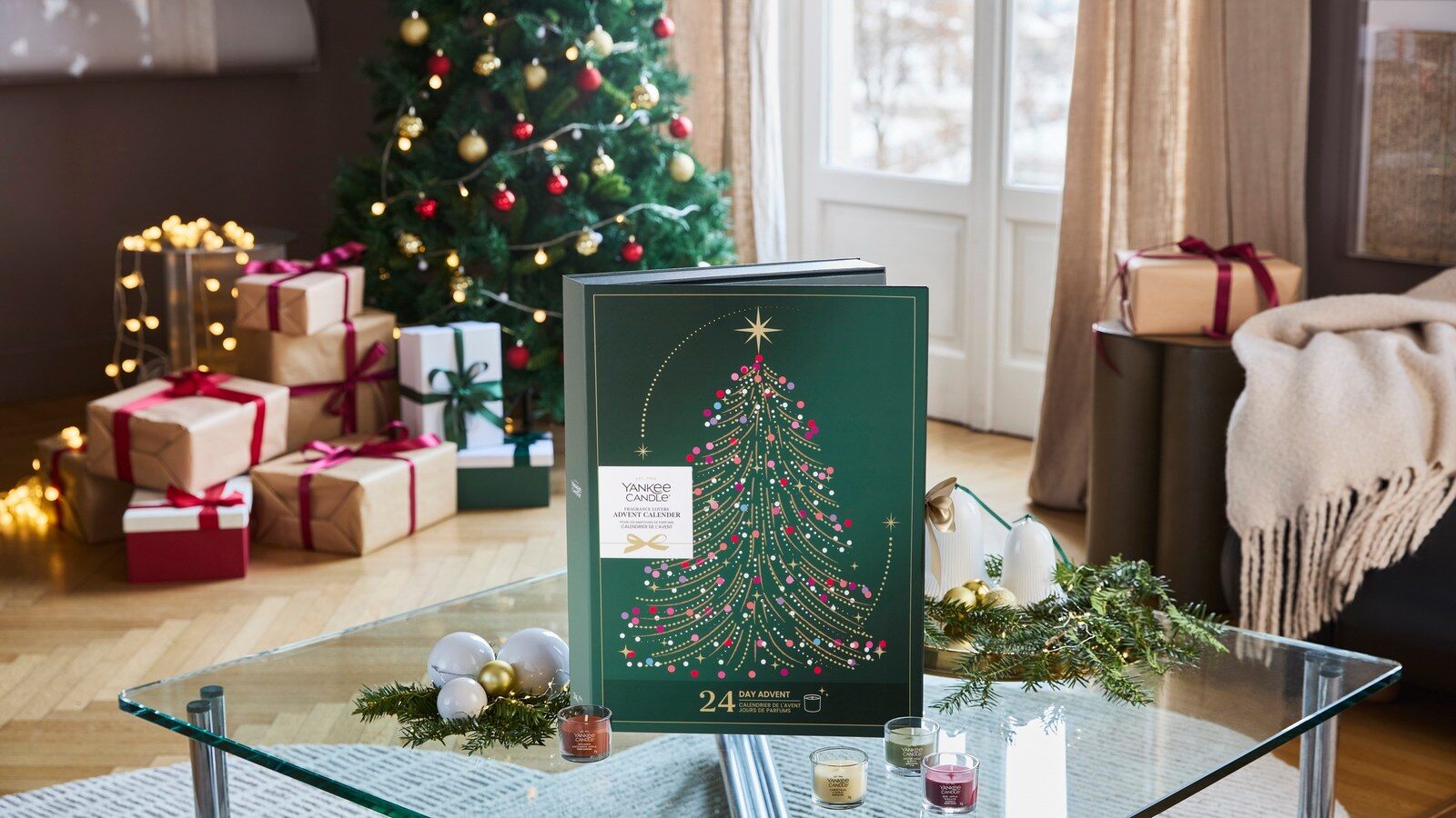 Le calendrier de l’Avent amateurs de parfums Yankee Candle posé sur une table basse en verre devant un sapin de Noël et des cadeaux