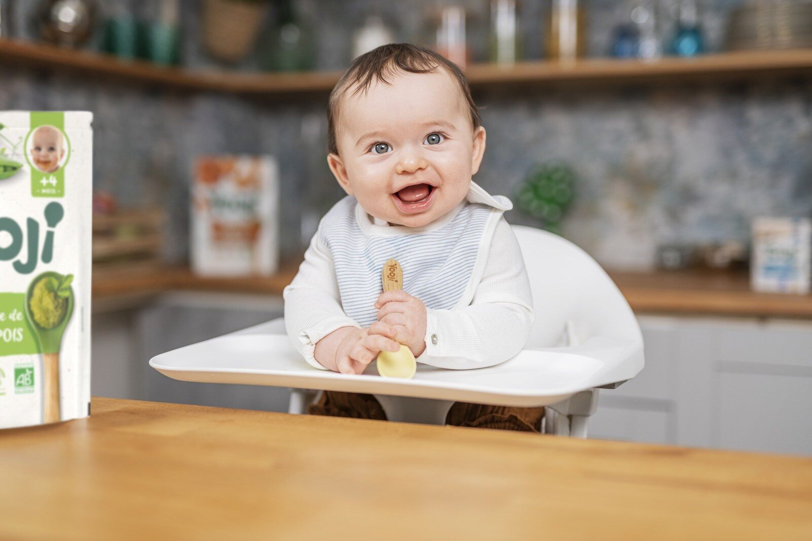 Un bébé souriant dans sa chaise haute porte une cuillère à sa bouche, devant une table de cuisine en bois et un sachet de purée Yooji