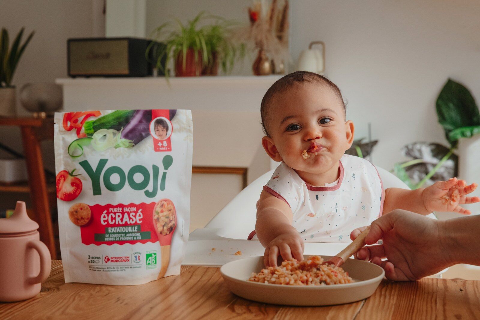 Un bébé assis à table mange un écrasé Yooji dans une assiette, devant un sachet de la marque et une plante
